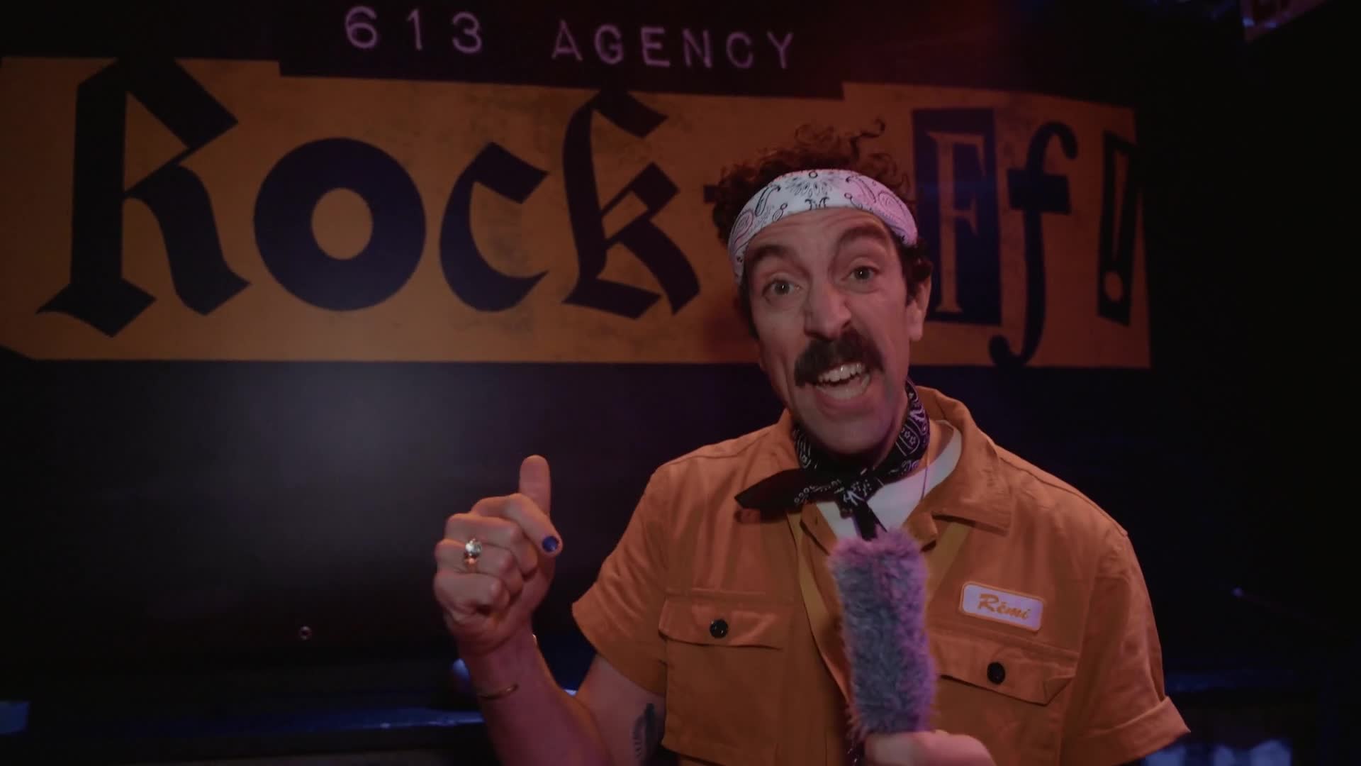 A host in a bandana and orange work shirt speaking into a fuzzy microphone in front of the 613 Agency Rock-Off banner